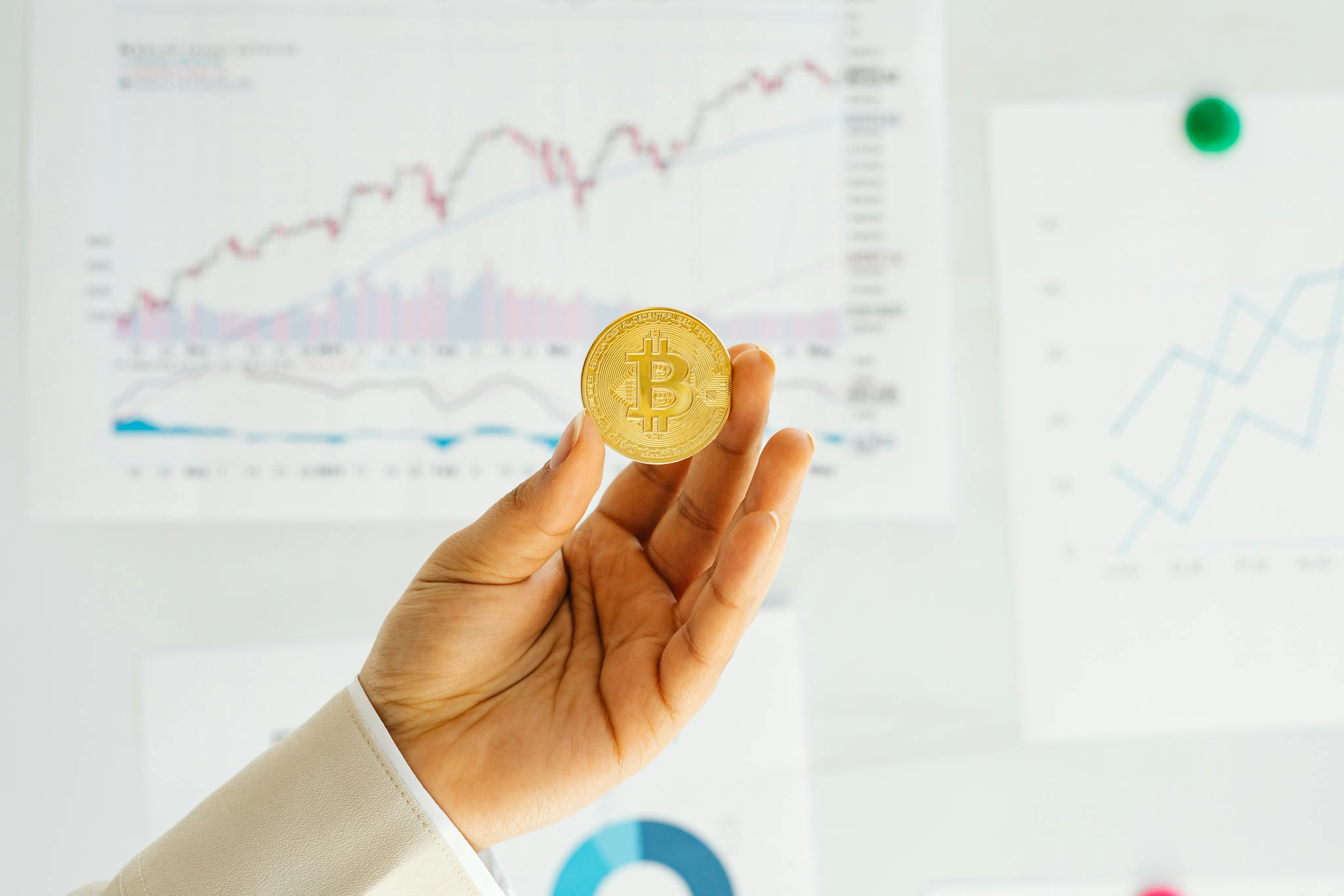 A hand holding a Bitcoin coin against financial charts depicting market trends.